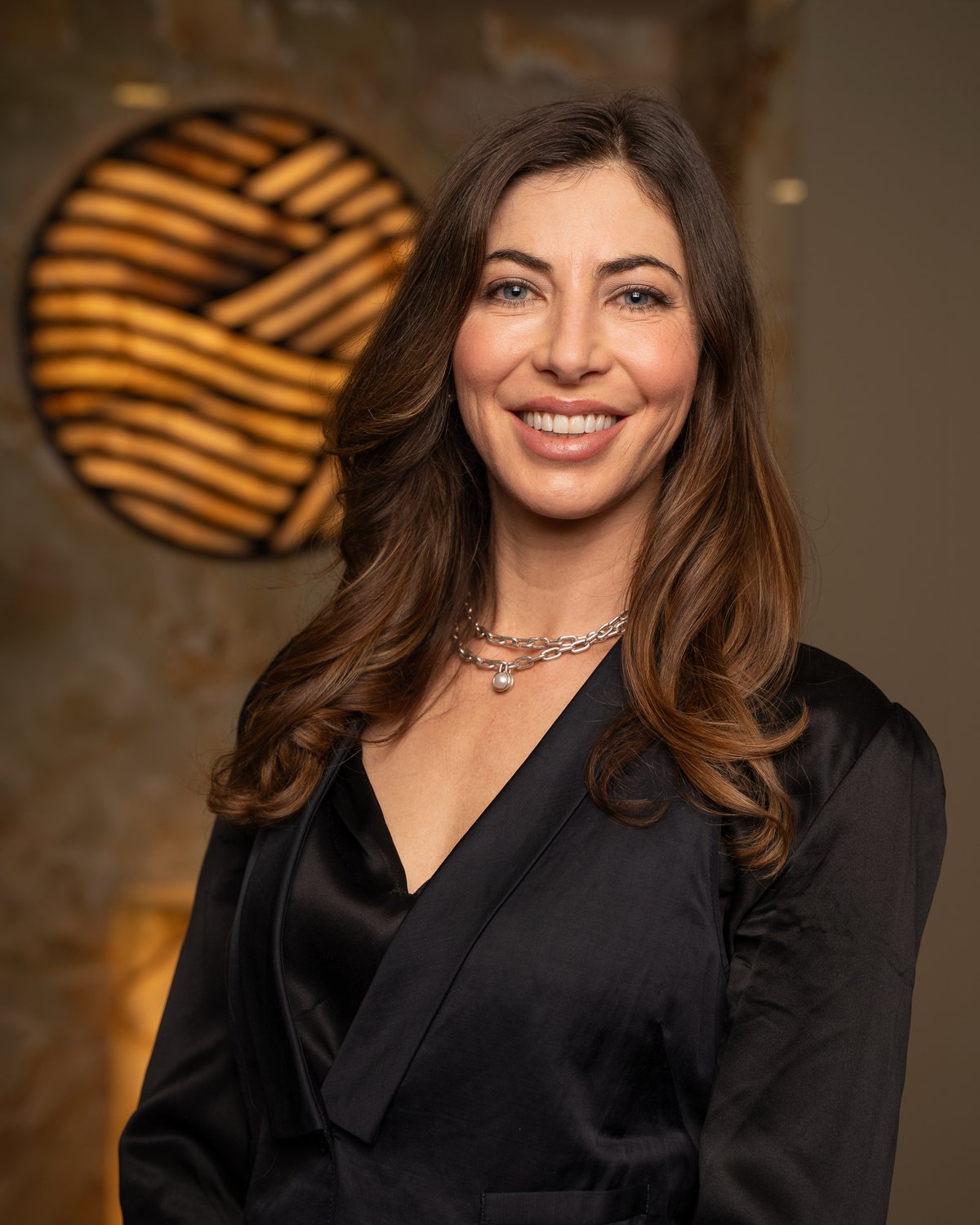 Dr. Sonia Bennett smiling in front of the illuminated emblem wall at Refined