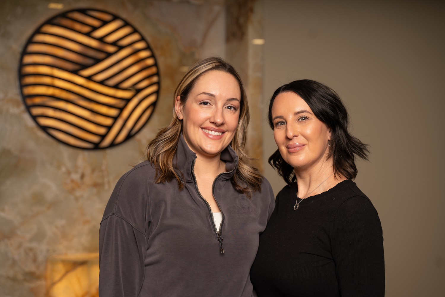 Two members of the Refined team smiling together in front of the illuminated emblem wall