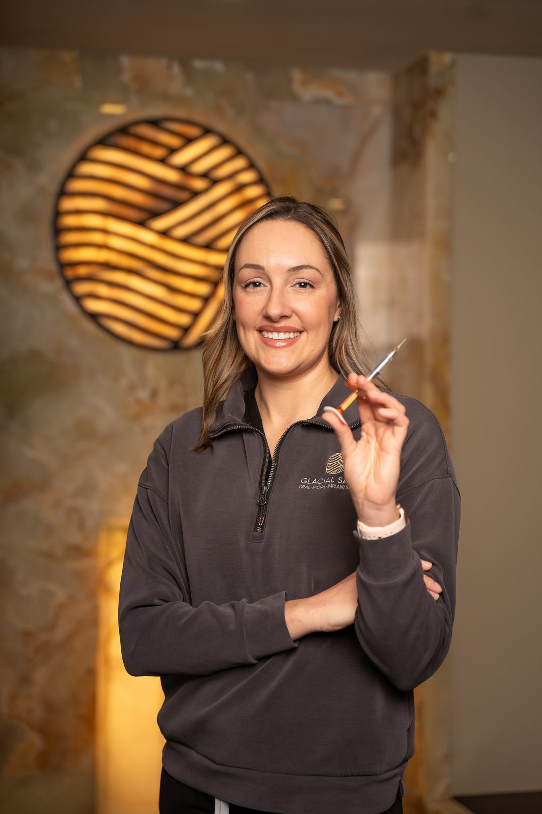 Refined's nurse injector smiling and holding a syringe in front of the illuminated emblem wall